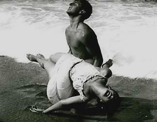 A scene of the film, black and white. A man is by the sea carrying a woman, who has apparently fainted or drowned. She is wearing a sleeveless, white dress.