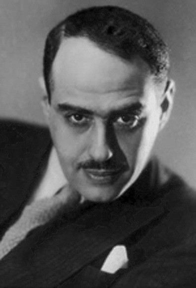 Black and white photo, undated. We can see a man with short, straight, dark hair. He has a black moustache and is wearing a black suit, a gray tie and a white shirt.