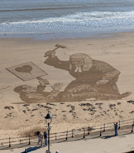 Sand art on the beach in Scarborough, United Kingdom of Banksy’s  lastest censored art depicting a judge with a gavel looming over a smaller figure holding a balloon drawing. Visitors admire the amazing work of art in the sand.