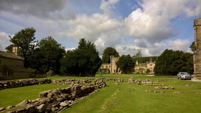 Amid the rolling greenery of Bolton Abbey in Skipton, remnants of ancient stone walls scatter across a neatly kept lawn, whispering of centuries-old history. To the left, a quaint stone building with a chimney sits quietly behind a low fence, while in the center, the grand façade of the abbey’s gatehouse rises with its arched entrance framed by tall trees. Sunlight breaks through a mix of drifting clouds, illuminating the golden stone of the historic structure against the deep green of surrounding woodland. In the distance, manicured grounds stretch toward the forested hills, evoking a serene balance of natural beauty and heritage.