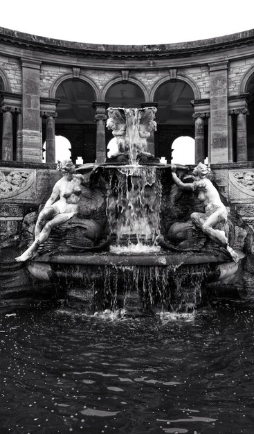 A dramatic, black and white, eye-level shot captures a grand classical stone fountain and colonnade. In the foreground, a dark pool of water, flecked with foam, reflects the scene. The ornate fountain features a central column from which water cascades forcefully, creating a dramatic, blurred effect. Two large, classical nude figures, carved from stone, recline on either side of the fountain's base. Behind the fountain, a semi-circular colonnade with multiple arches and pillars frames the upper part of the image, showcasing intricate architectural details and leading the eye upwards. The overall mood is timeless and imposing, enhanced by the monochrome palette.