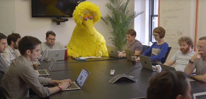 Big Bird (a Sesame Street character that is a large, yellow bird) sitting at one end of a conference table, drawing with crayons on a pad of paper, surrounded by a group of young human (not bird) comedy writers, all with laptops open in front of them.