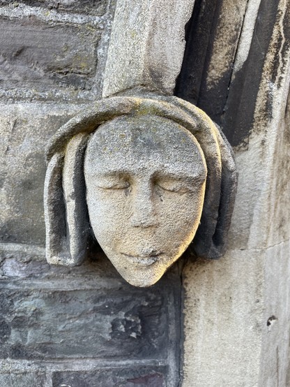 A carved woman's face with a set mouth and closed eyes, wearing a maybe medieval headscarf 