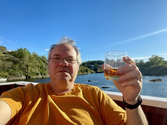 A selfie of a pale man nearing 60, wearing an orange t-shirt and holding a glass of single malt scotch. In the background is water, some hills and vegetation and a clear blue sky.