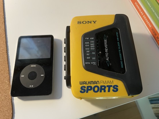 On the left: a black iPod with click wheel. From sometime around 2003-04. On the right: a yellow Sony AM/FM Sports Walkman. Perhaps from 1986-ish.