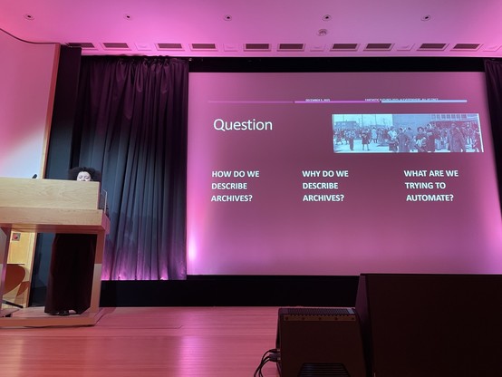 A light-skinned Black woman is just visible behind a podium on a stage. Her slides ask:

How do we describe archives?
Why do we describe archives?
What are we trying to automate?