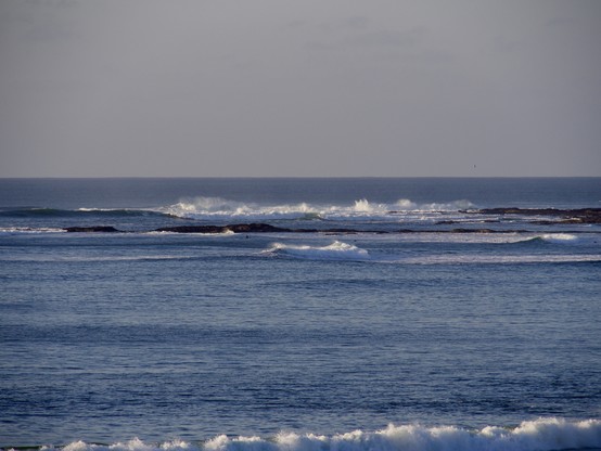 Waves lapping on the shoals, throwing white spray into the air. The sky is clear and blue.