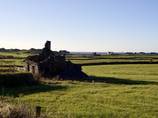 A ruined croft in a green field. In the background is Spanish Point golf club, and some houses. Beyond them is the ocean. The sky is perfectly clear and blue.