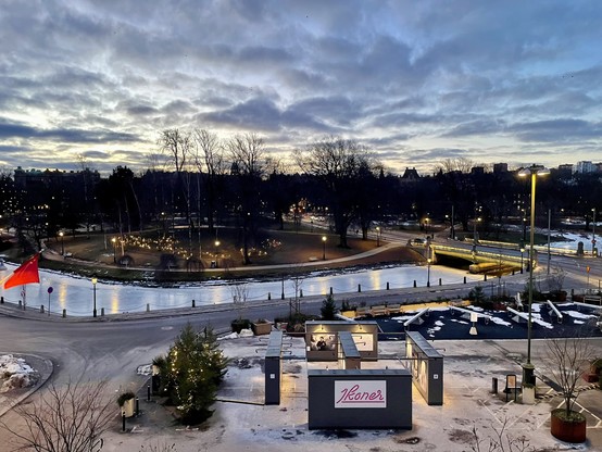 A view of Gothenburg from a hotel window. In the foreground is a small square. Behind it is the moat that once went around the walled city, the water in it frozen solid. Beyond the moat is a park filled with winter trees, and the tops of buildings can be seen beyond them again. Finally, the pre-dawn light fills the eastern sky which is dotted with light clouds.