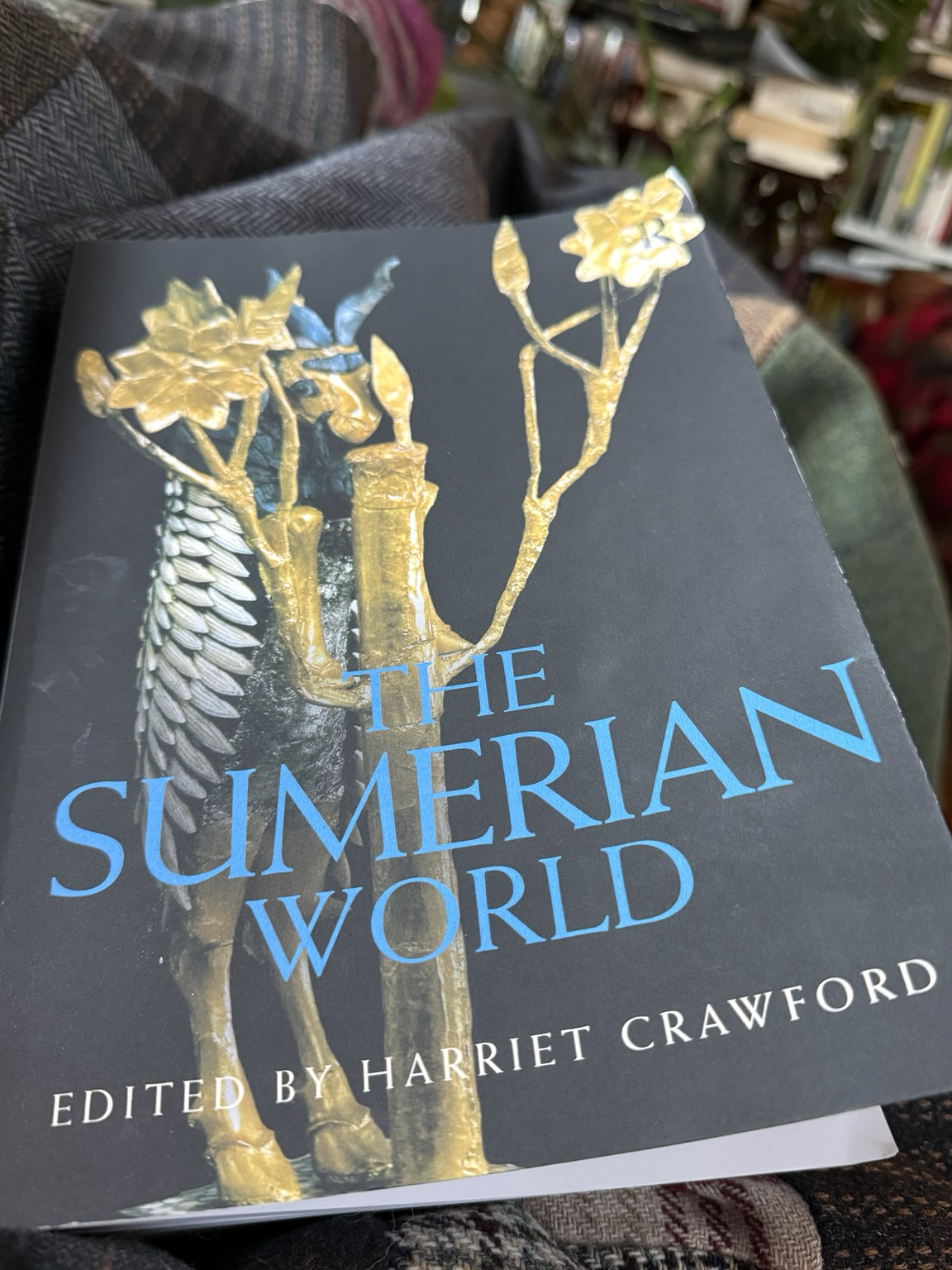 Cover of The Sumerian World edited by Harriet Crawford. Title in blue on a black background, though showing a gold ornament of a cool, but shy-looking goat (?) with a very cool feathered coat, standing on its hind legs behind a golden tree —I am guessing somehow related to Asherah. 