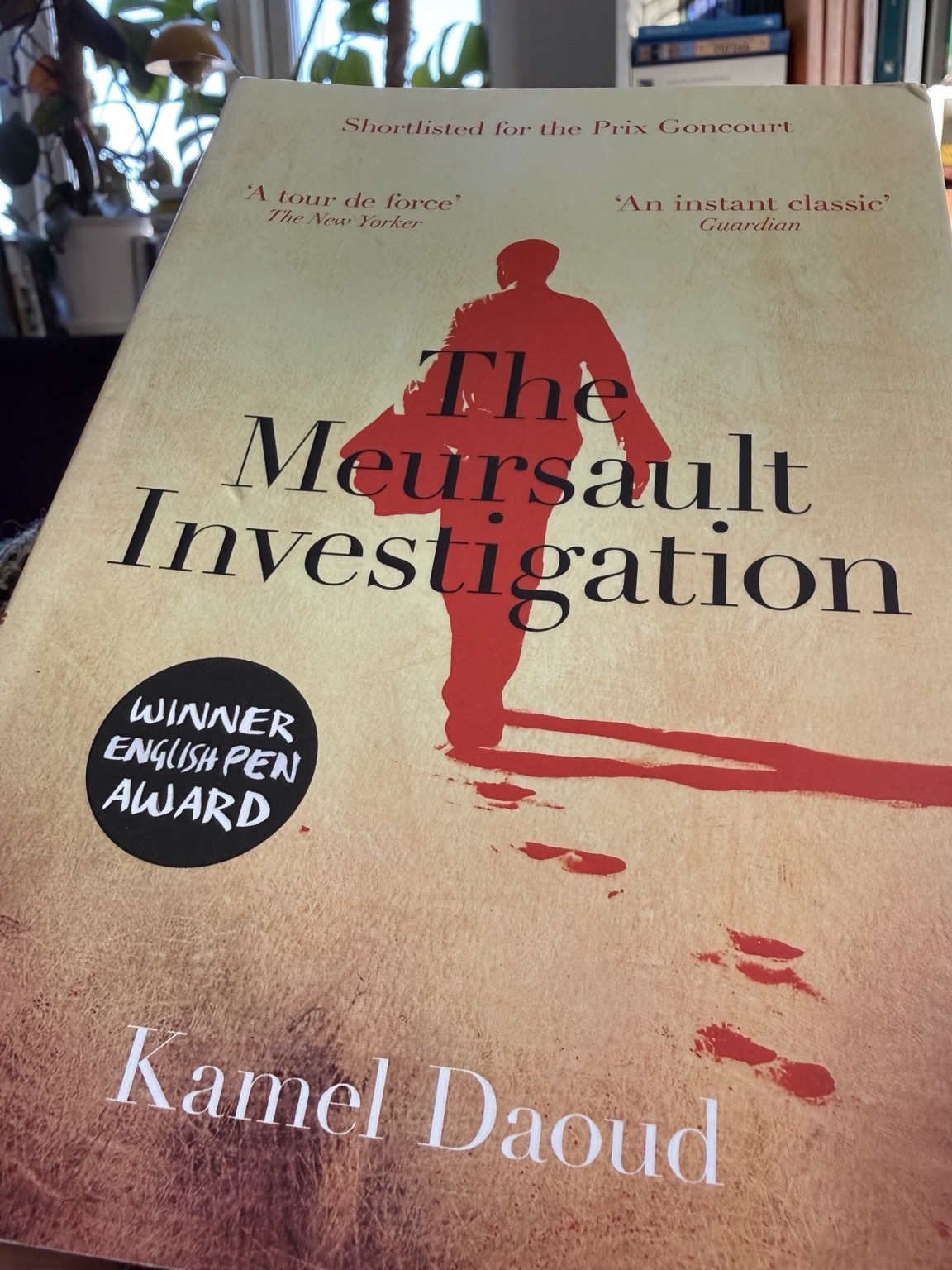 Cover of The Meursault Investigation. A red burnt-out figure walks over sand on the cover.