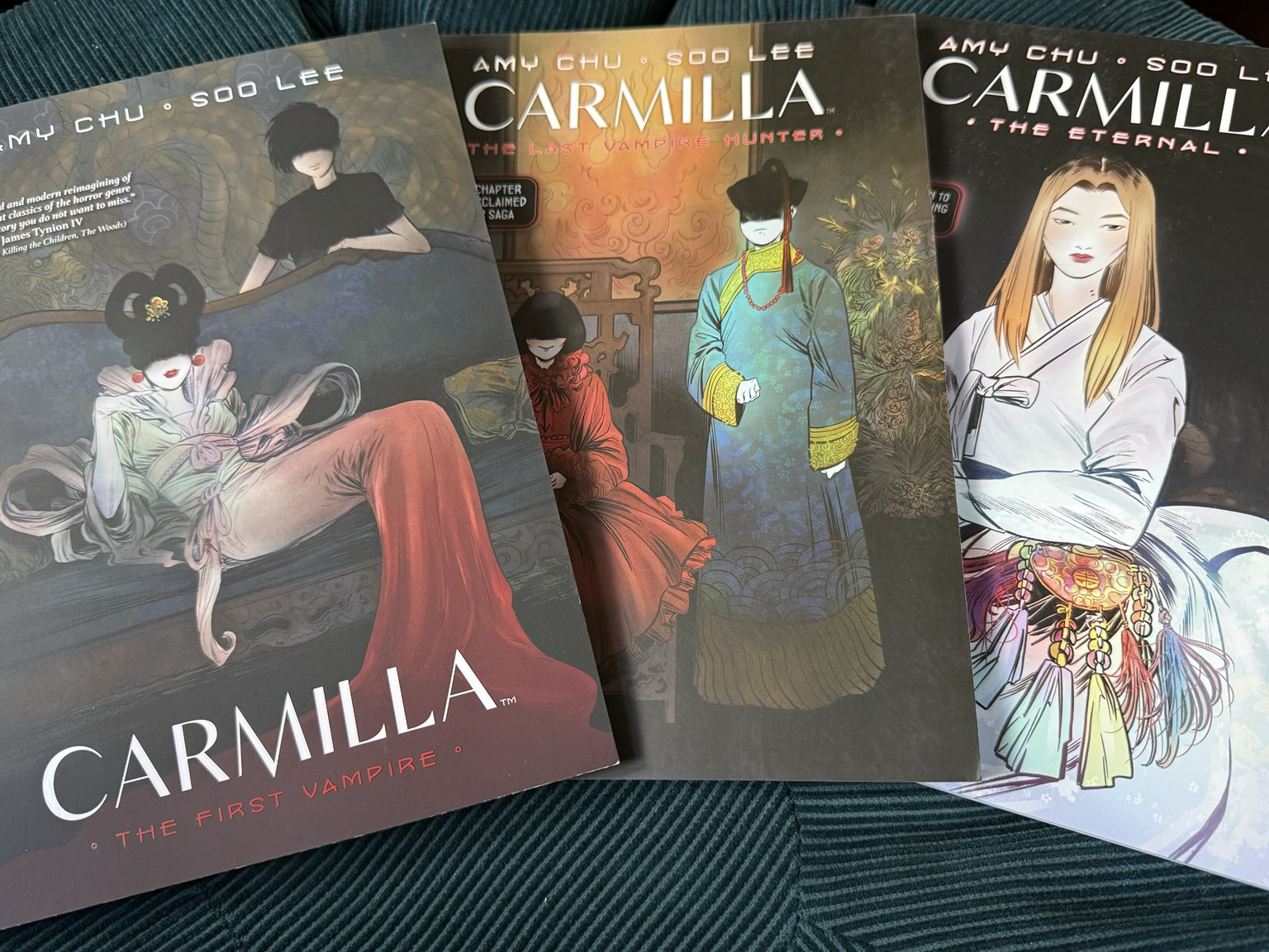 Photo of the three TPs of Carmilla: The First Vampire, The Last Vampire Hunter, and The Eternal. The first two covers are in earthy and dark jewel tones, one showing a woman in a mix between Chinese and French dress lounging on a sofa while another in a dark t-shirt stands behind. Both their faces are obscured. The same is true of the two figures of the second cover, where the character who is fully visible wears ancient Chinese clothing. The third cover shows a young woman whose face is not obscured. She wears a white, probably Chinese, possibly Korean dress against a dark background, with a colourful bag.