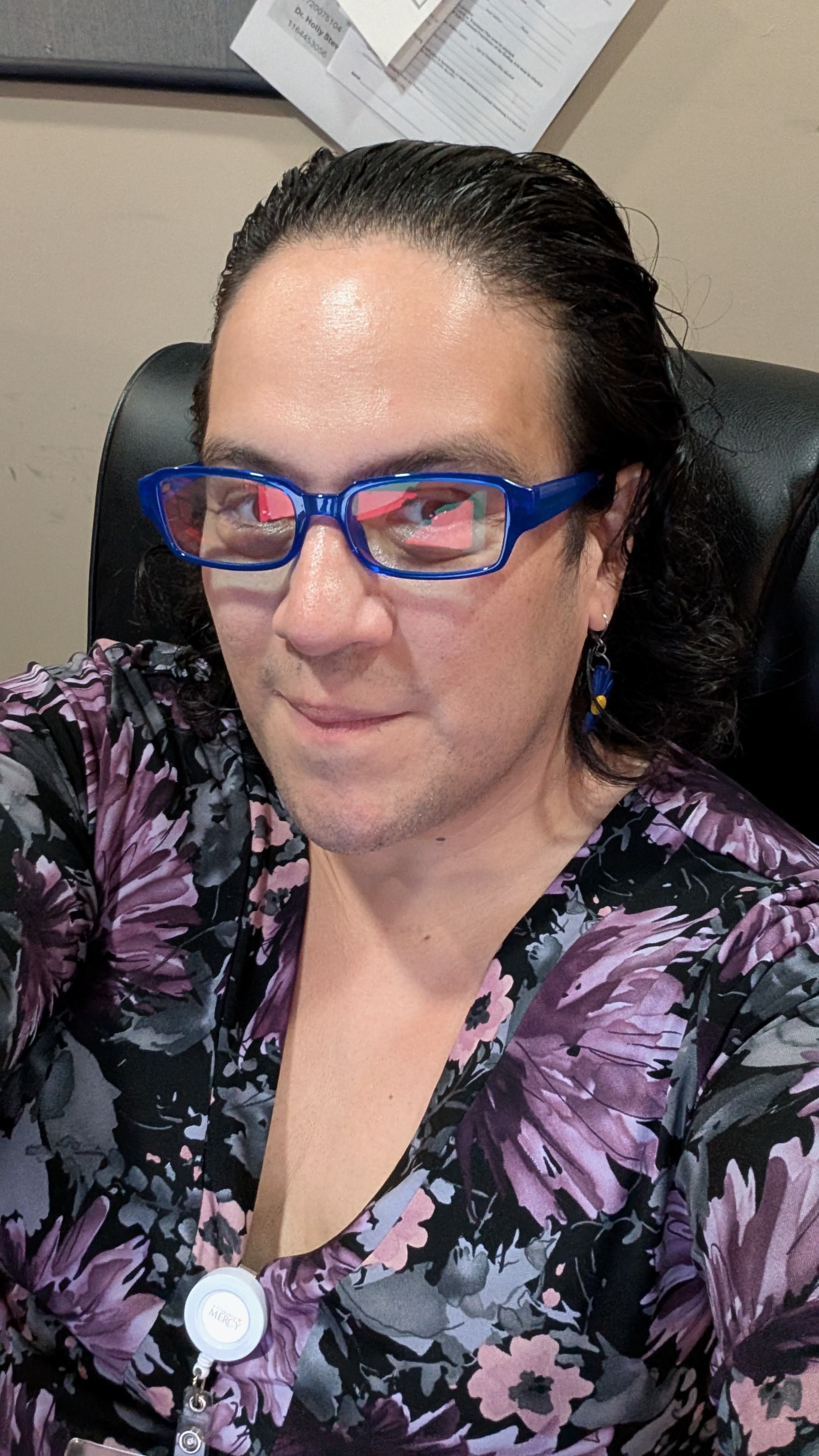 A close-up selfie of Ellis seated in a black leather office chair. She is wearing a black v-neck top with a vibrant purple, pink, and grey floral pattern. She has dark, slicked-back hair, bright blue rectangular glasses reflecting a digital screen, and a dangling earring featuring blue and yellow elements. Clipped to the neckline of her top is a white retractable badge reel displaying the Pittsburgh Mercy logo. The background consists of a beige wall and a dark-framed bulletin board displaying pinned papers, one of which contains printed text and numbers.