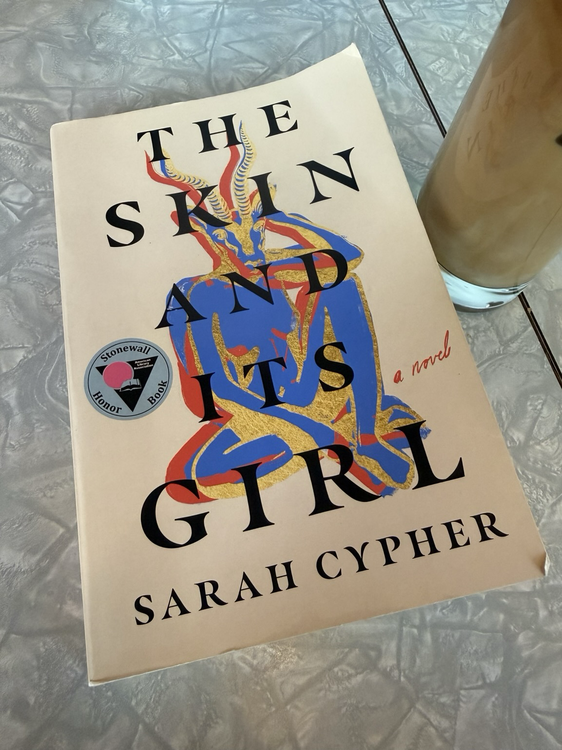 The book on a grey table next to a coffee. The cover is cream with black lettering, over a painting of what looks like a female satyr/devil figure in blue red and gold. The book is marked with a circle featuring a triangle with a book and a pink dot and text noting it as the American Library Associations Stonewall Honor Book.