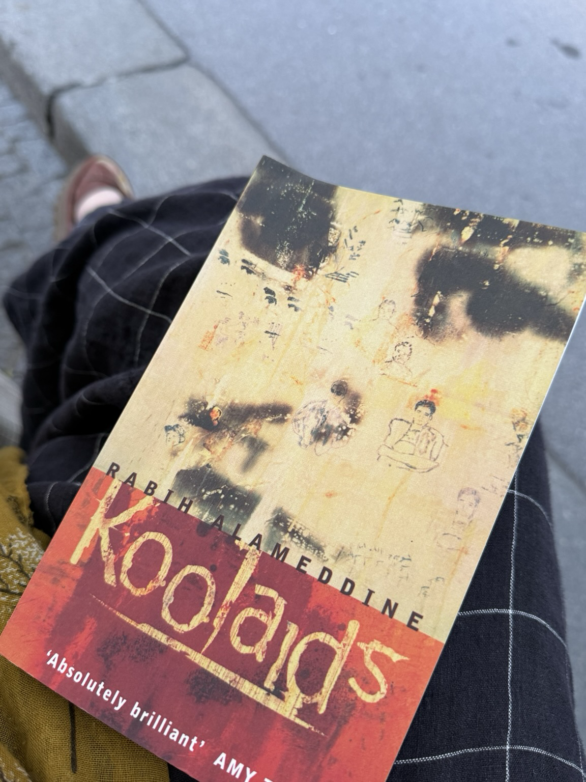 Cover of Koolaids against my knee and a curb. Cover shows title in yellow over a red block in bottom third. The rest is a blurred photo of a face looking perhaps worried? Sad? Small figures drawn across.