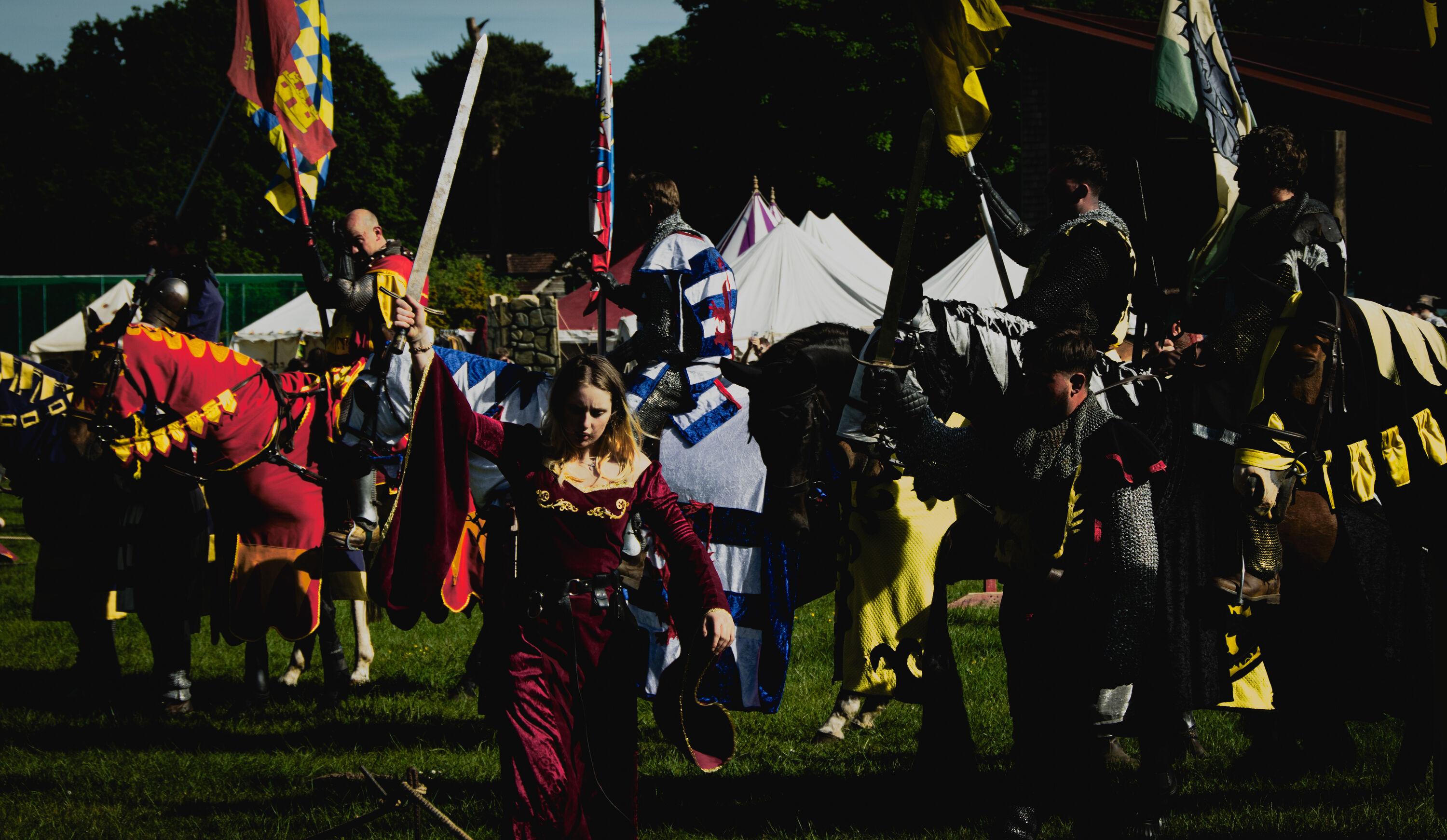 A line-up of medieval reenactors in costome: knights on horses and on foot carrying flags; a lady brandishing a sword.