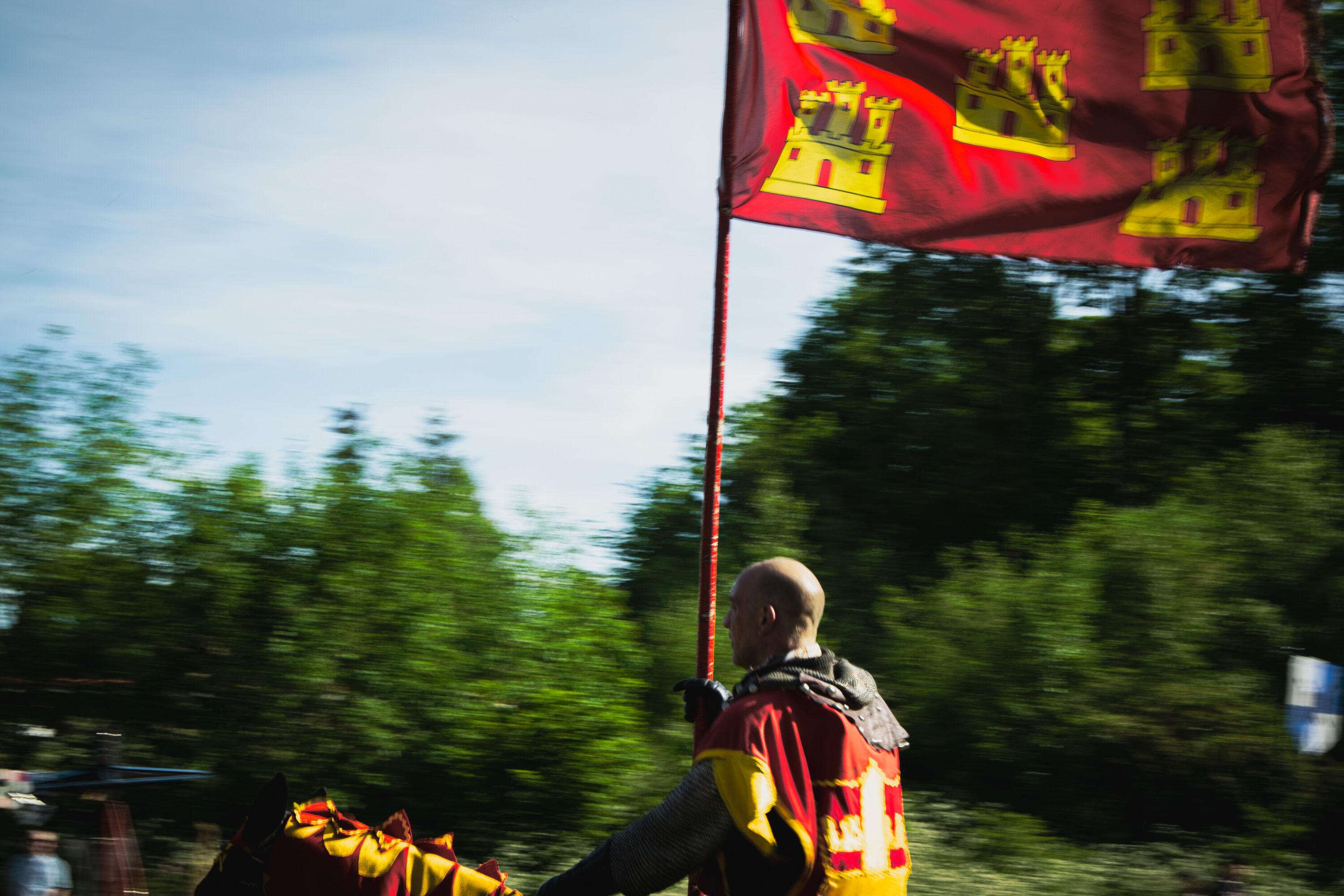 A reenactor dressed as medieval knight riding a horse, carrying a flag.