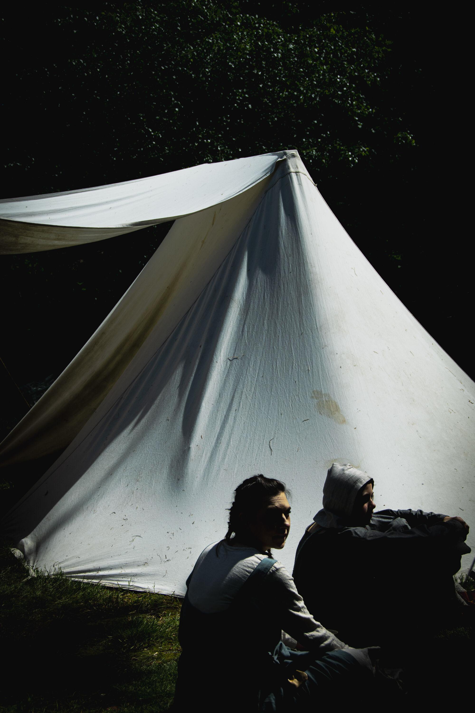 Reenactors: two women dressed up as medieval folk sitting in front of a white tent in the woods.
