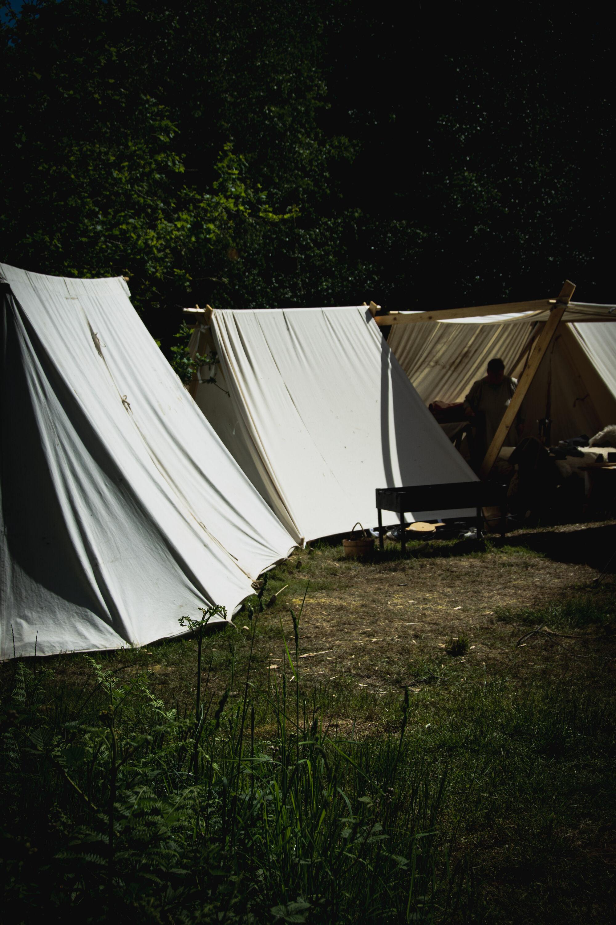 A group of white tents in the woods. A figure is faintly visibile in the shadows inside the last tent on the right.