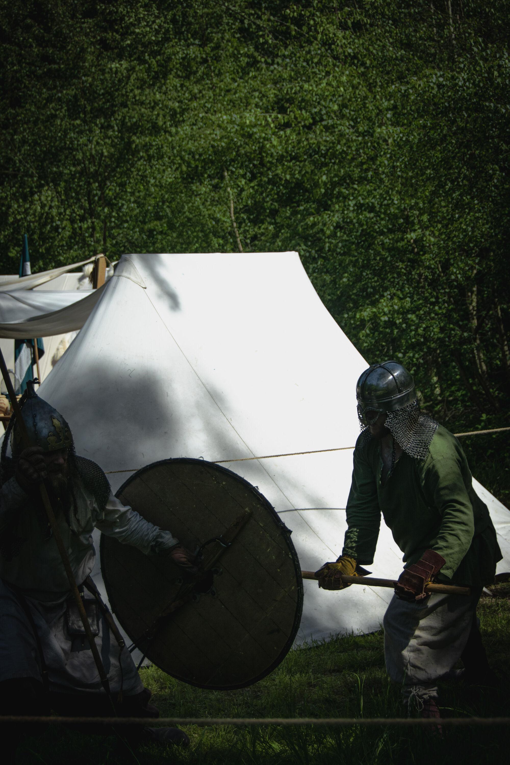 Two reenactors dressed as medieval warriors staging a mock fight; the figures are in the shadowed foreground against the bright, sunlit background of a white pitched tent.