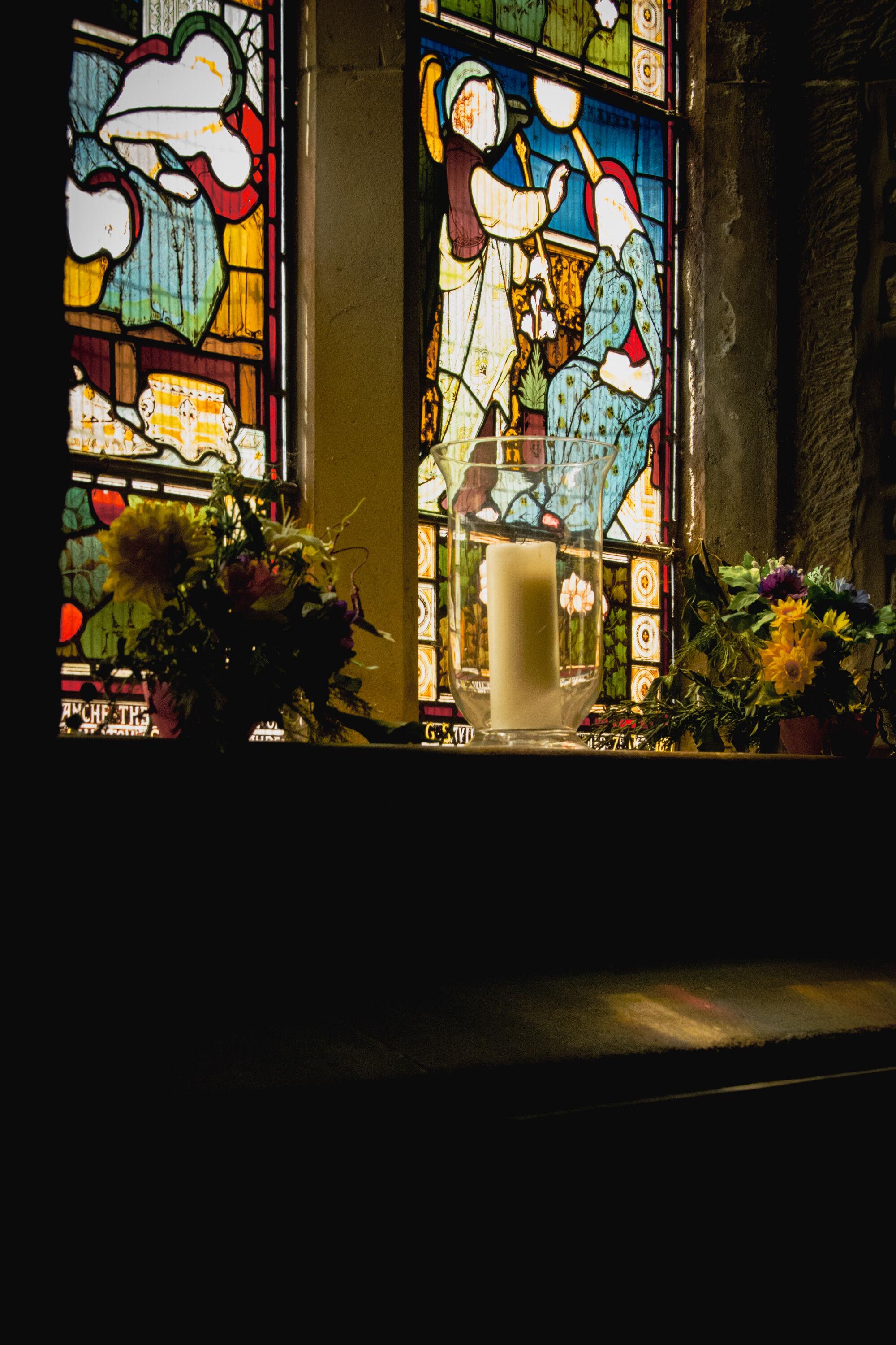 Inside a church, looking slightly upwards towards a stained-glass window, with a candle and flowers in front.