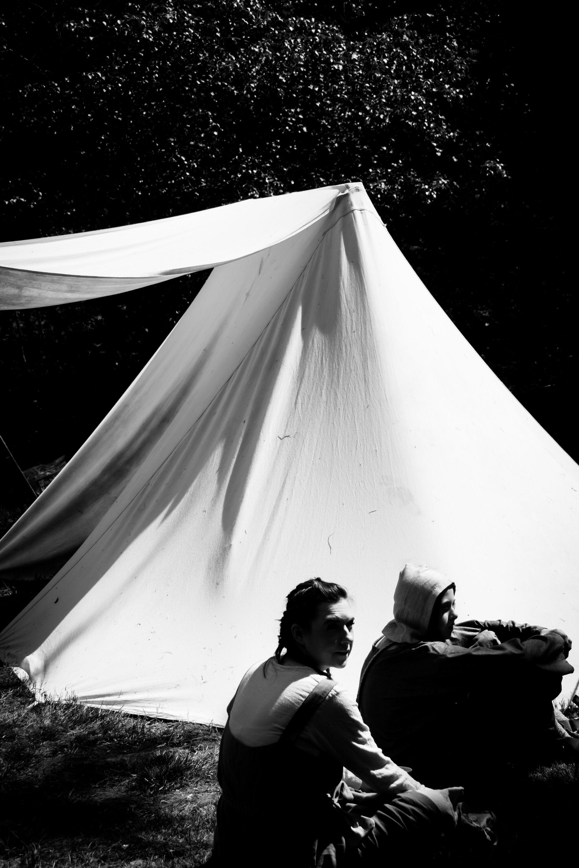 Two women dressed in medieval garb sitting in front of a pitched tent.  The image is black and white with strong contrast between light and shadow. It was taken during a Robin Hood themed reenactor event in early summer.