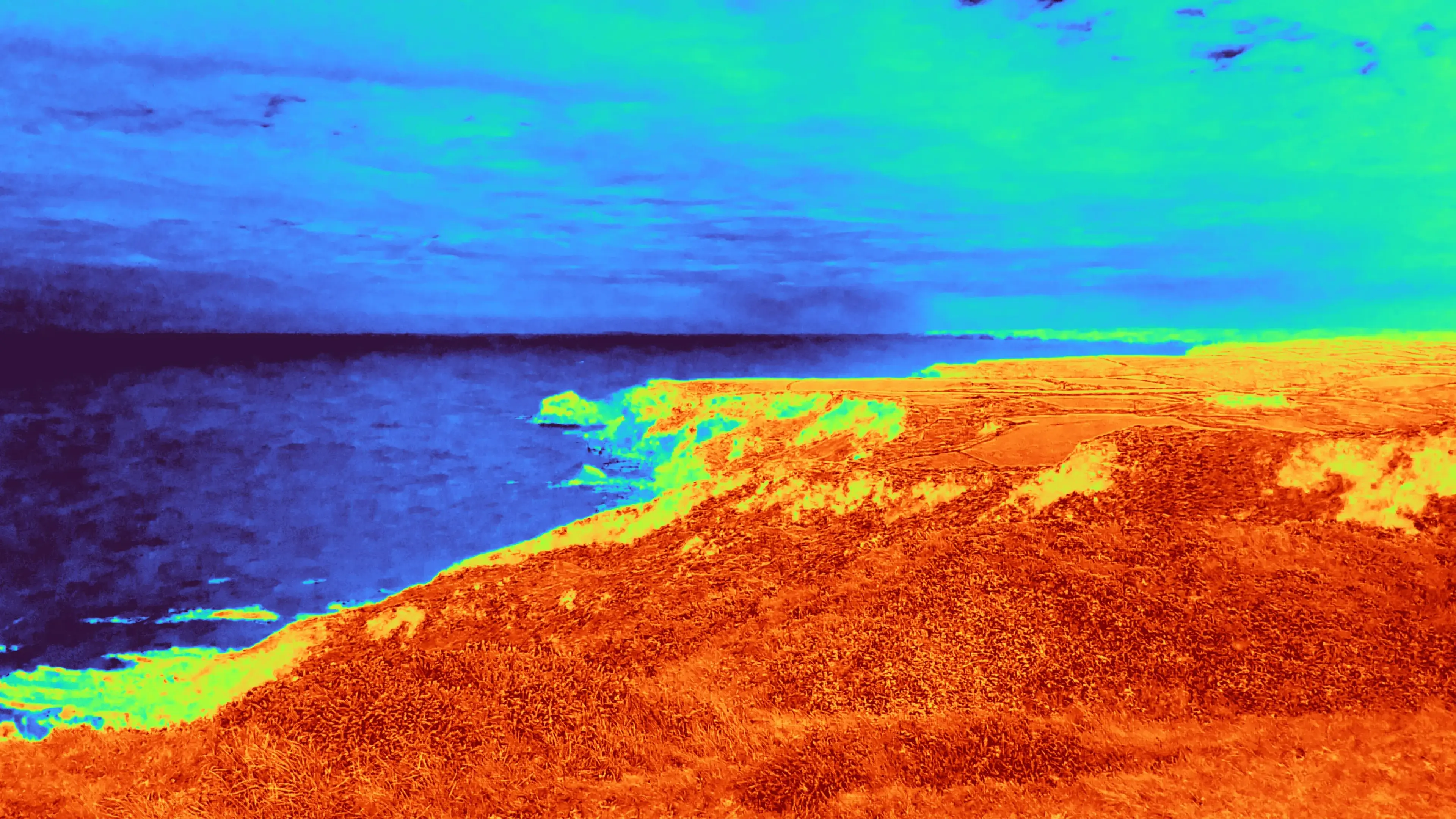 Vibrant false colour, NDVI indexed image of the same scene. All vegetation glows red, areas with scrubby vegetation, adhering to bare rock, are more yellow. The sea, sky, and coastal rocks are green to blue, indicating little absorbed moisture - that is, no vegetation