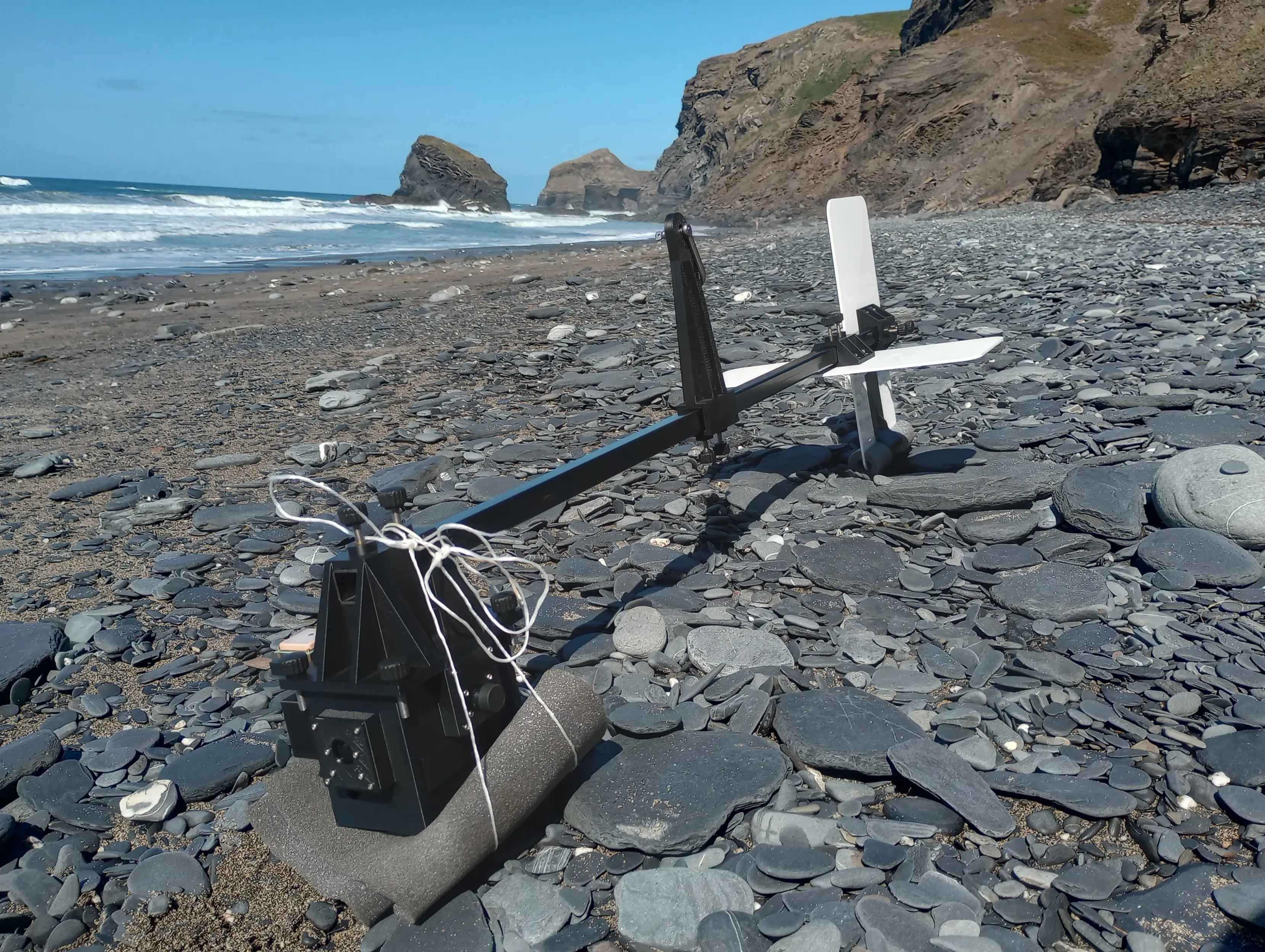 The Kite Drone rests on the stones of a beach, with the sea in the background, along with rugged cliffs, and a brilliant blue sky. It consists of a long central spar, with a sensor box on one end, two cruciform tale fins on the other, and a tall kite line mount in the centre.