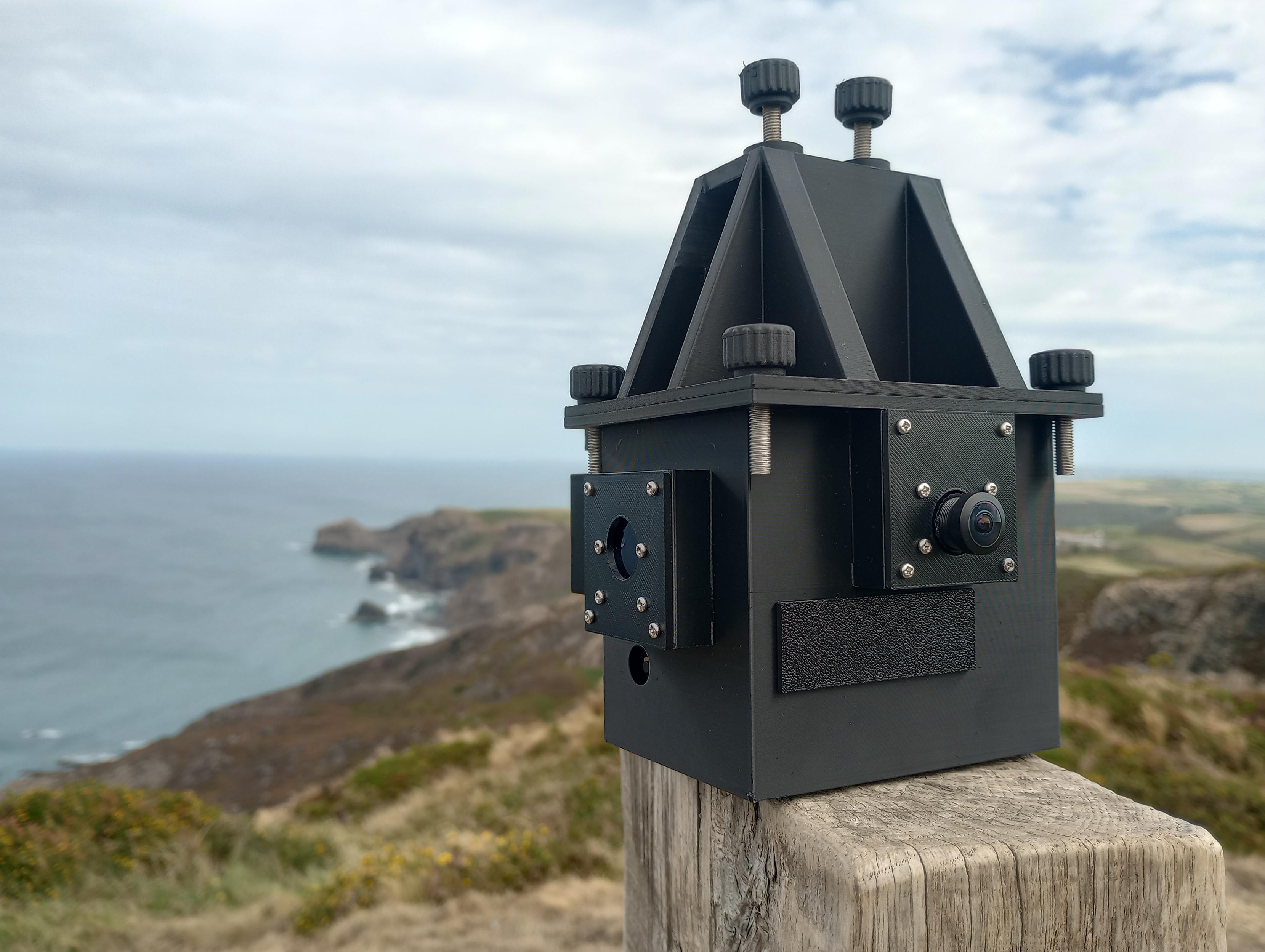 A black sensor box, with a couple of camera ports and a pyramid shaped attachment hat, sits on a fence post. In the distance is the beautiful north Cornish coastline.