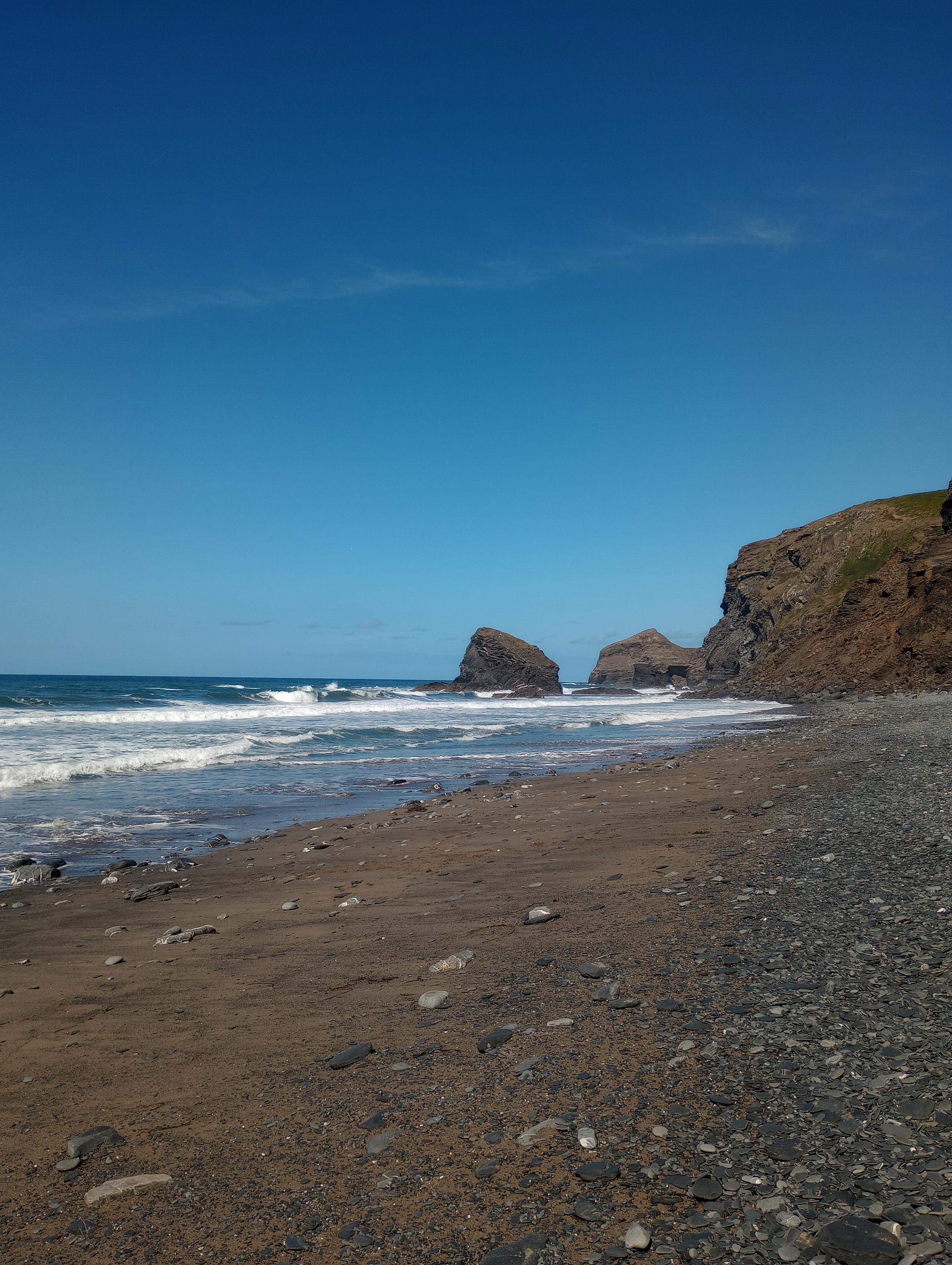 A rocky beach shoreline, with rugged cliffs in the background. The sky is a brilliant clear blue