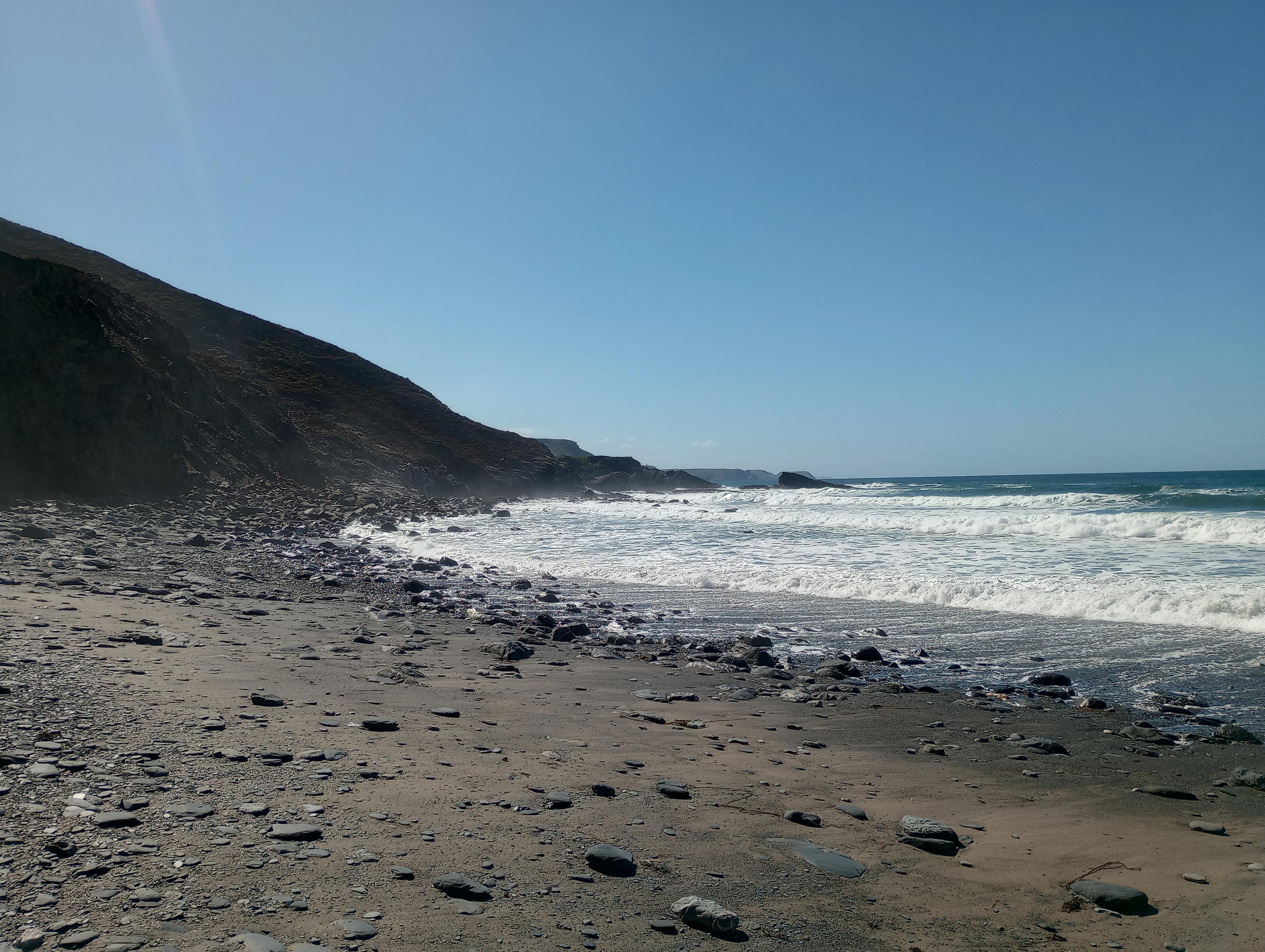 A rocky beach shoreline, with rugged cliffs in the background, taken from another angle. The sky is a brilliant clear blue