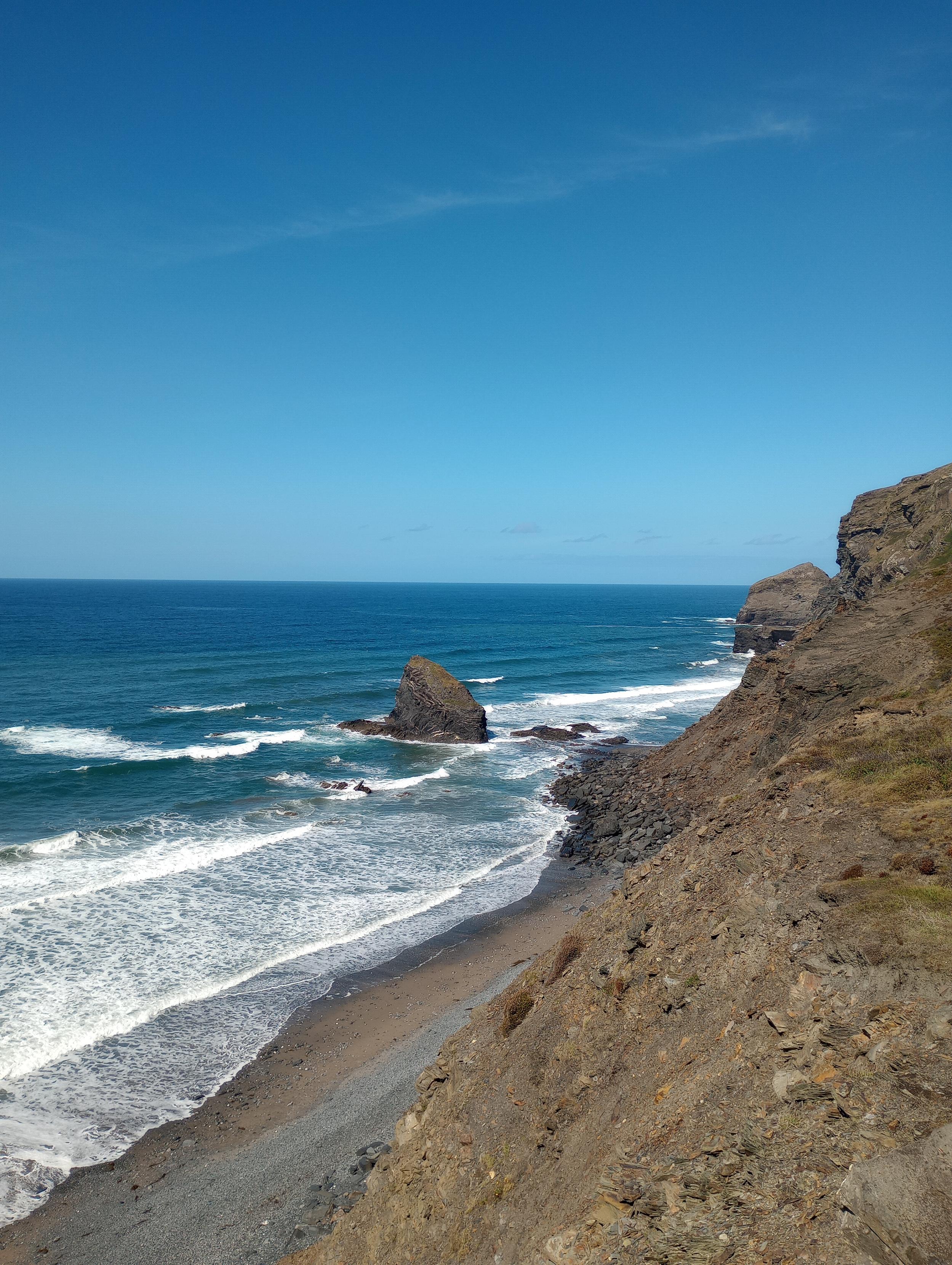 A rocky beach shoreline, taken on the descent down, with rugged cliffs all around. The sky and the sea are brilliant shades of electric blue